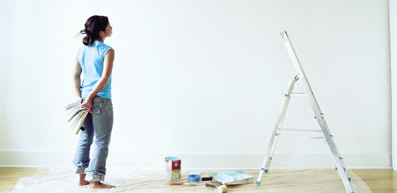 Woman holding paint swatches looking at white wall.