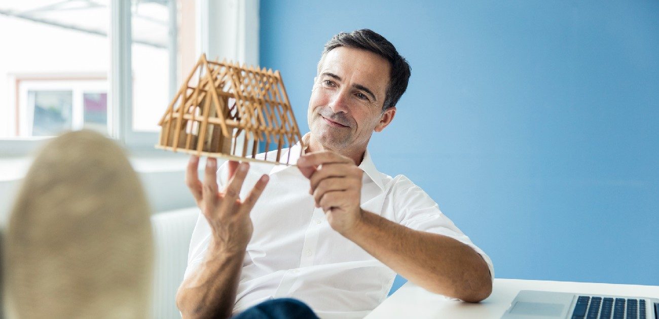 A man sitting at a desk in a home office holds a wooden-framed house miniature. 