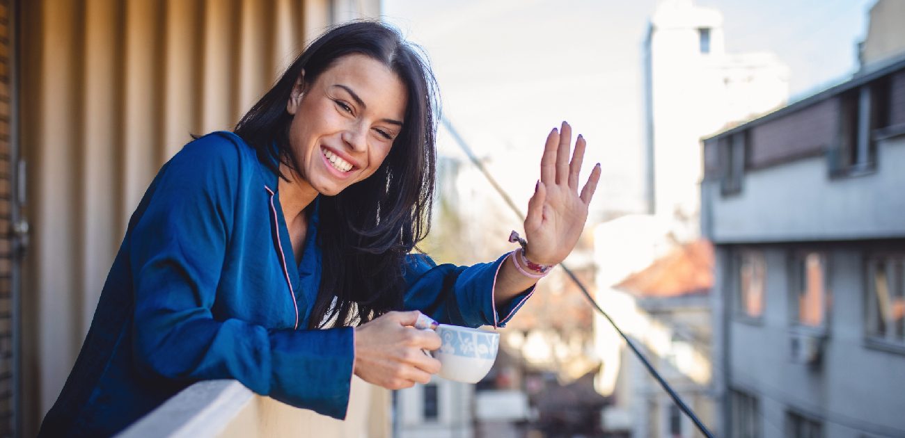 A smiling woman waves at the camera while drinking coffee on her balcony.