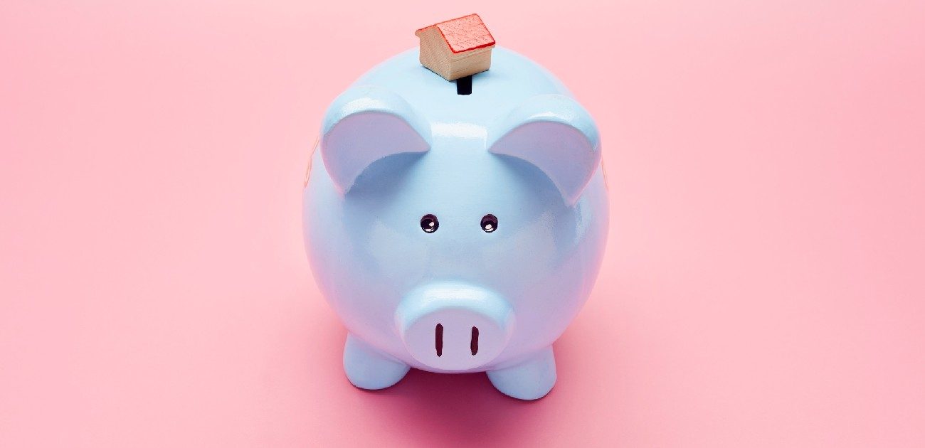 A picture of a light blue piggy bank with a tiny house miniature resting atop it, with a pink background.