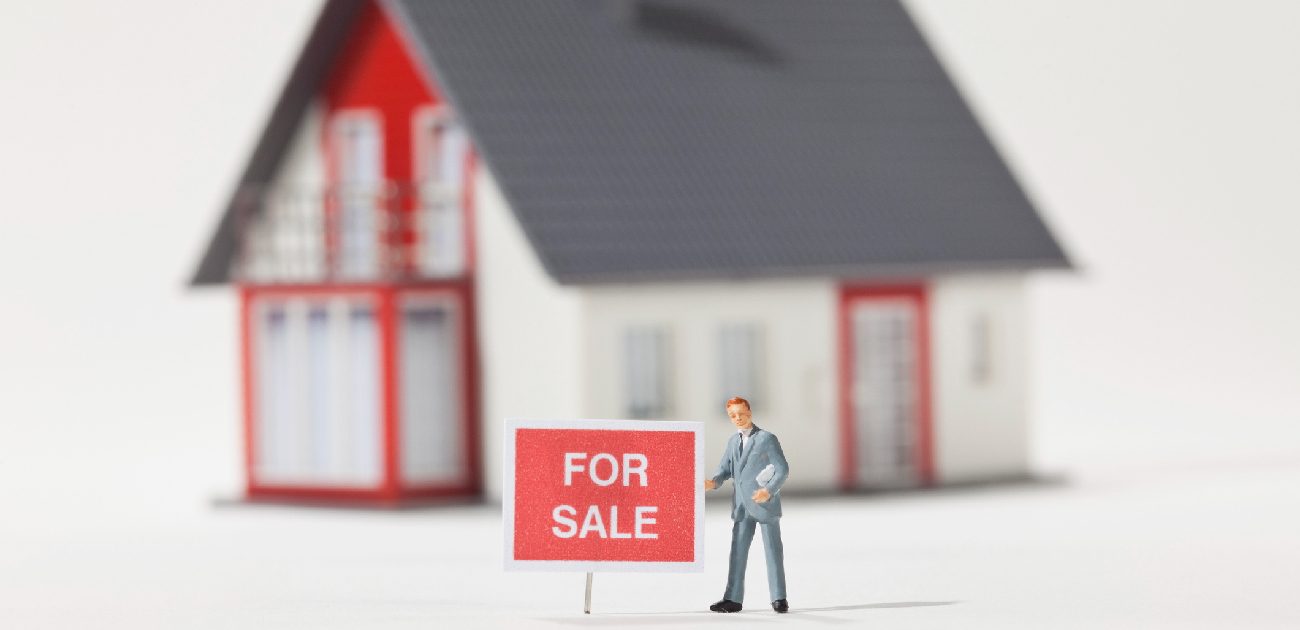 A graphic of a house on a white background with a For-Sale sign and a person in the foreground.