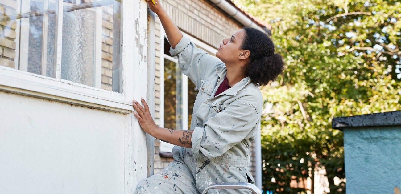 A woman assess chipping paint on the corner of an enclosed porch’s exterior wall.