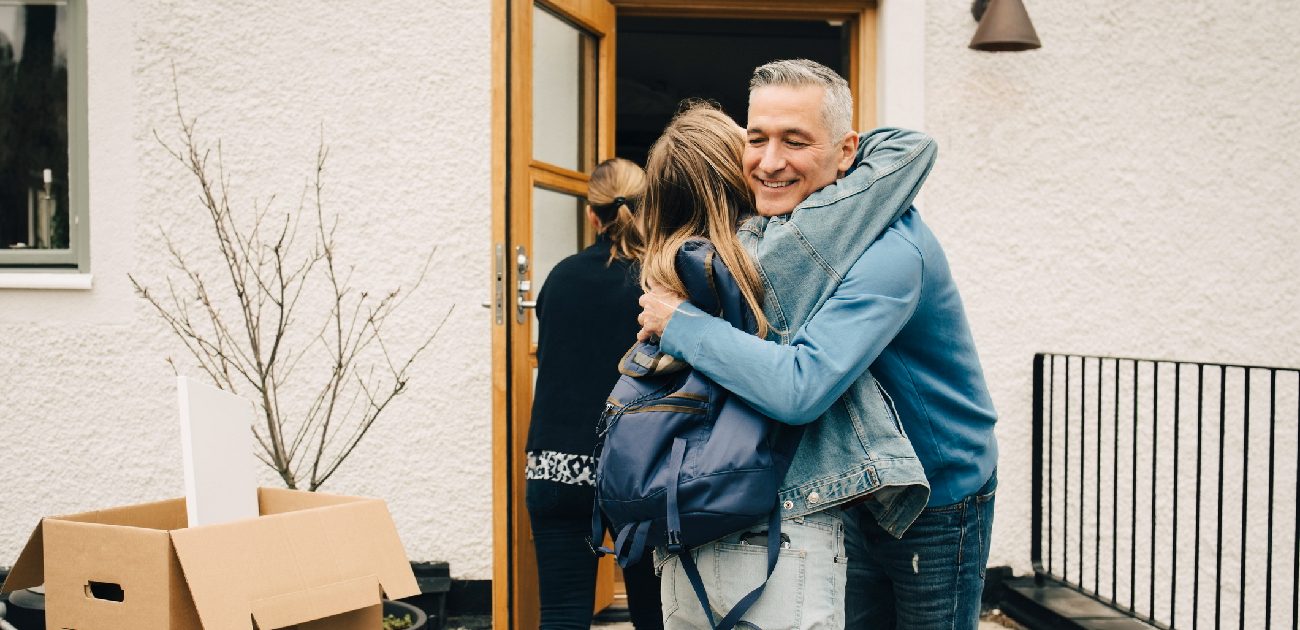 A father and daughter hug at the front door of their home as she moves back in, with stacked boxes to their left on the ground.