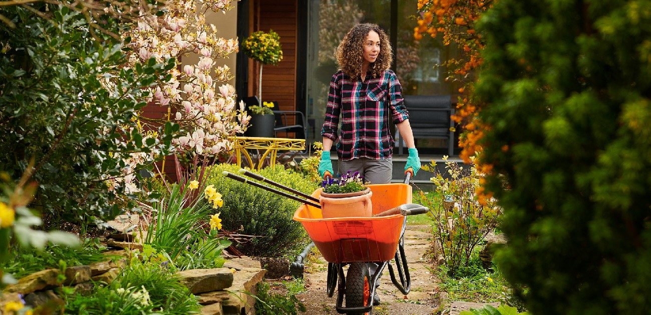 A woman pushes a wheelbarrow while working in her yard, with flowers and plants all around.