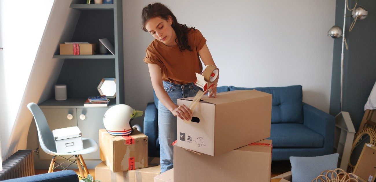 Picture of a woman in her living room taping up a moving box stacked upon other ones.