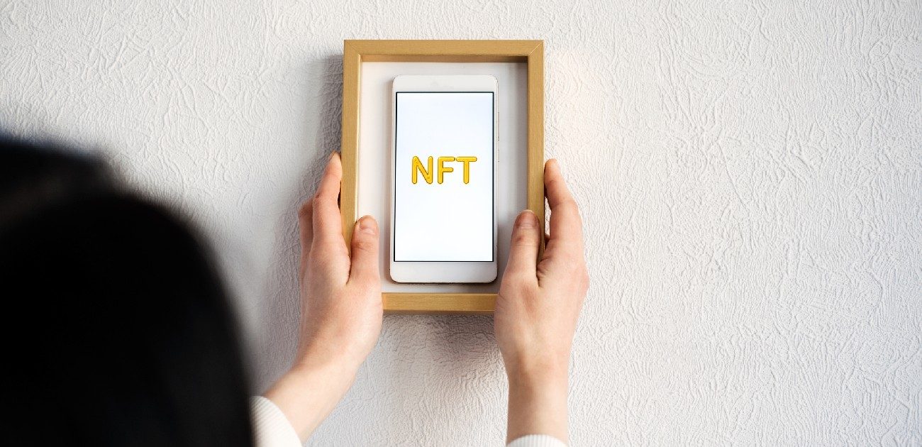 A person holds a framed smartphone displaying the term "NFT" on its screen.