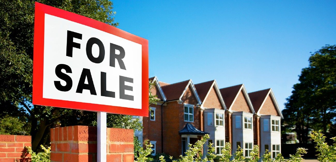 A picture of a For Sale sign in daytime standing before a row of houses in the background.