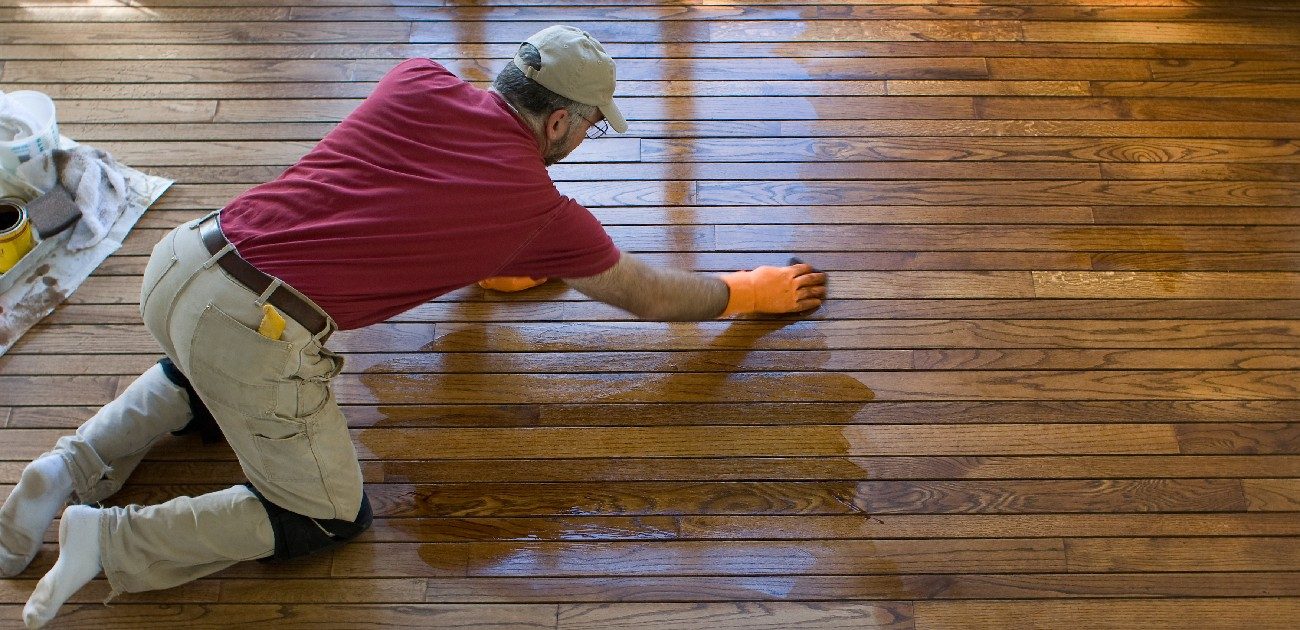 A home renovator on his hands and knees on a hardwood floor adding polish to it.