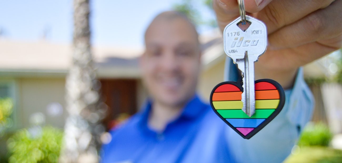 A smiling man holding a house key attached to a fob shaped like a heart with a rainbow.