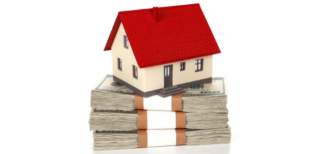 An image of a house miniature sitting upon three thick bound stacks of money on a white background
