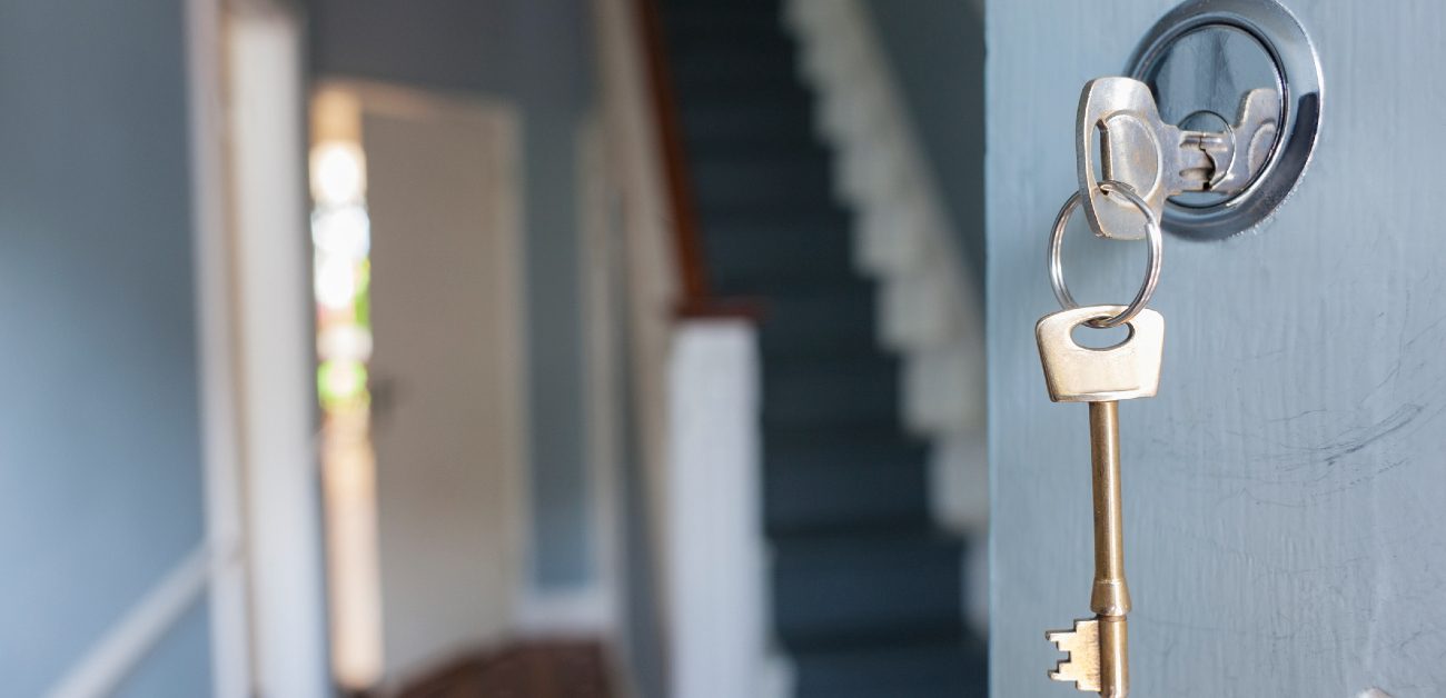 A picture of a set of keys in the front door lock of a new home that is partly open, showing the foyer and staircase.