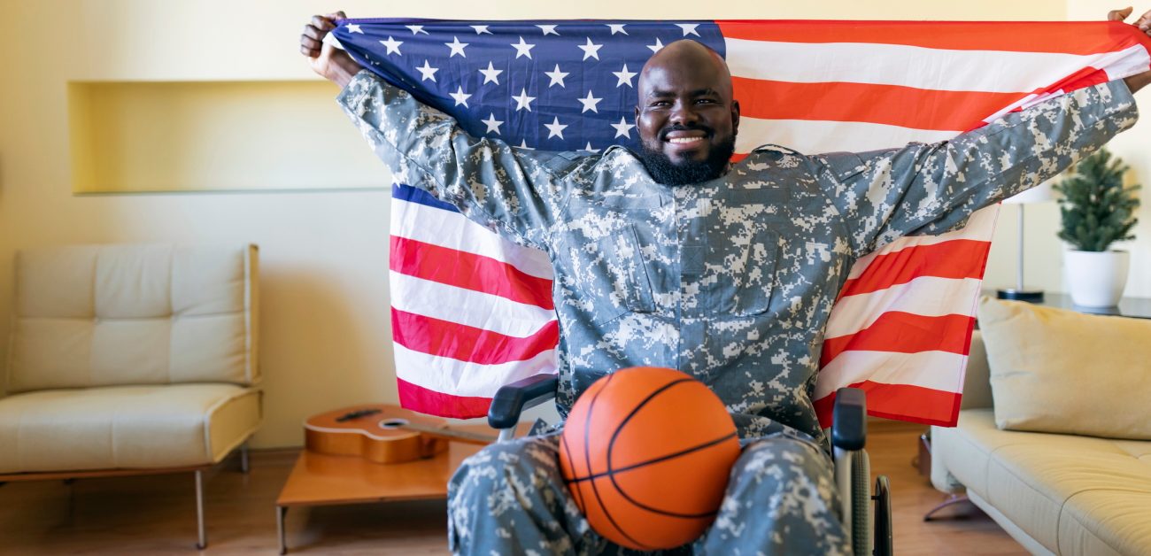 A grinning Black veteran in a wheelchair holds up an American flag in his home