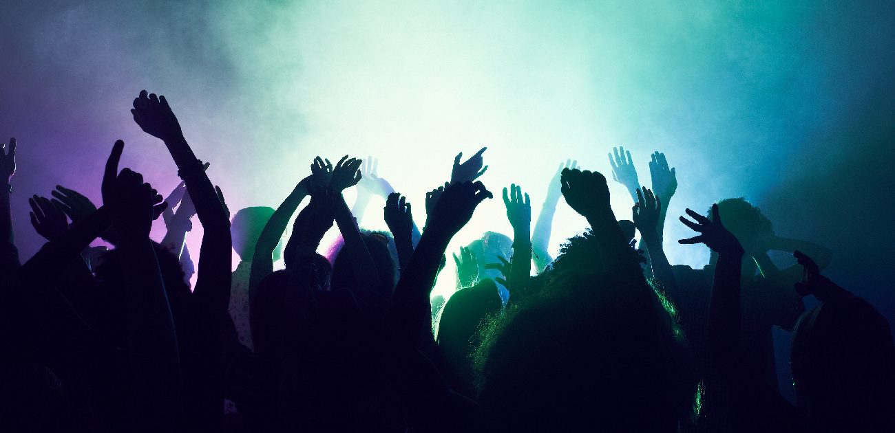A silhouetted crowd of concertgoers with their hands in the air, with blue-green and purple lighting and fog.