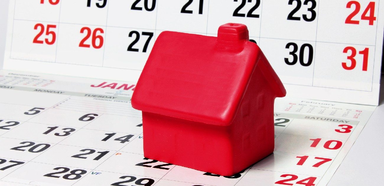 A small red house miniature like a Monopoly house is sitting on a calendar, with another calendar standing vertically behind it.
