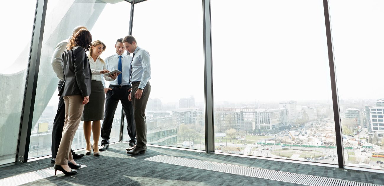 A group of coworkers in a high-rise office building stand near a wall of windows overlooking the city.