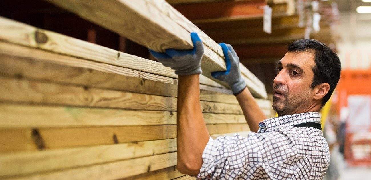 A man at a lumber yard picks up two large two-by-fours from a stack of the same cut.