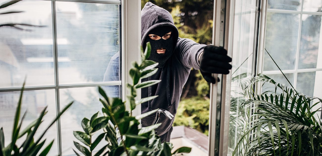 A burglar donning a ski mask and black gloves entering a home through tall retractable windows.