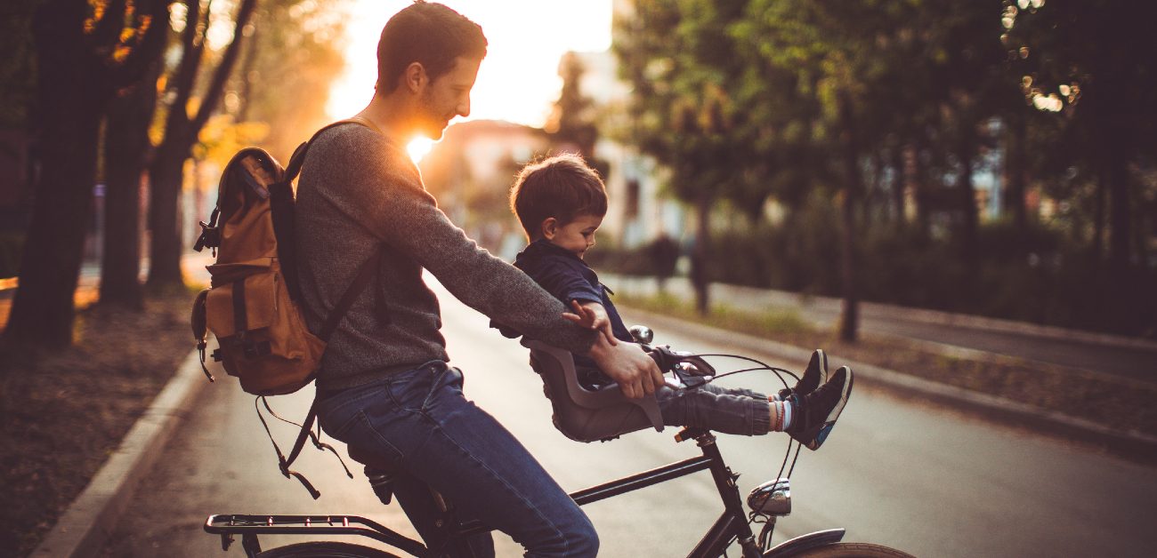 A man rides his bike at sunset across a street, holding his son steady on the handlebars.