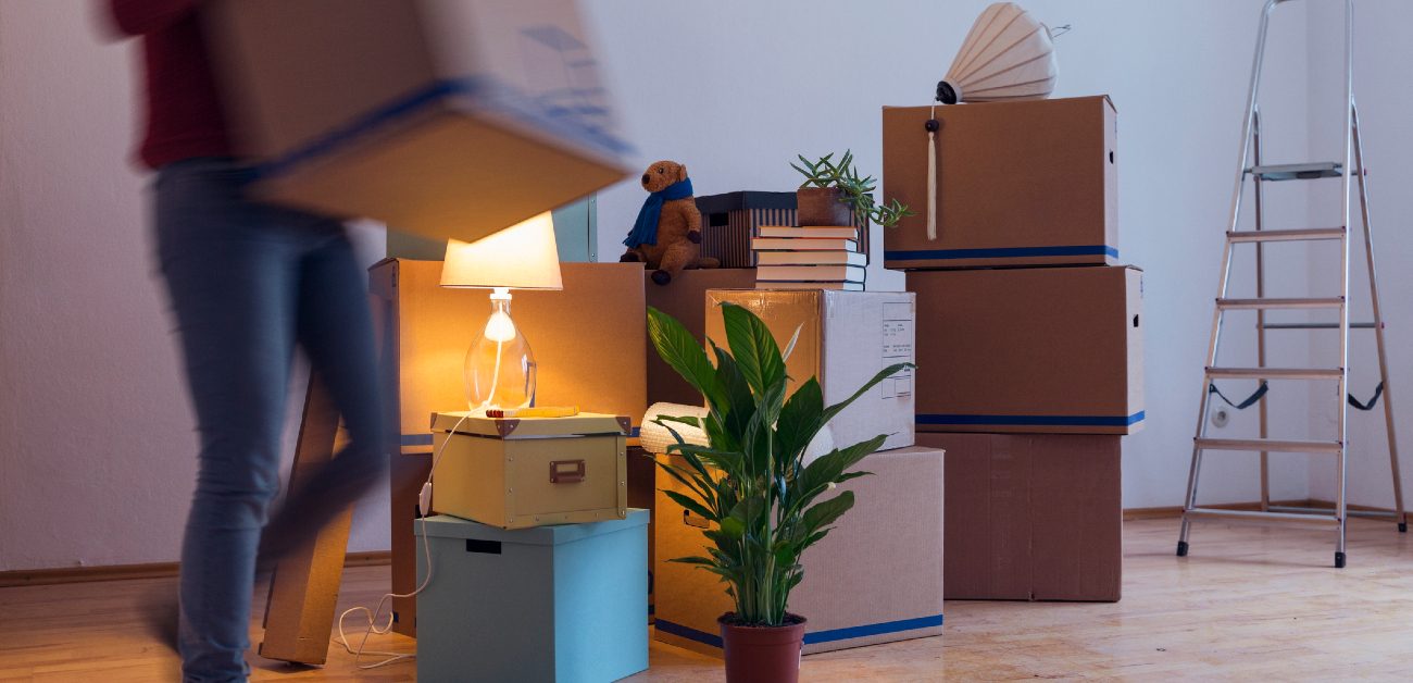 A picture of a person carrying a box in an empty room with other boxes and belongings stacked up together.
