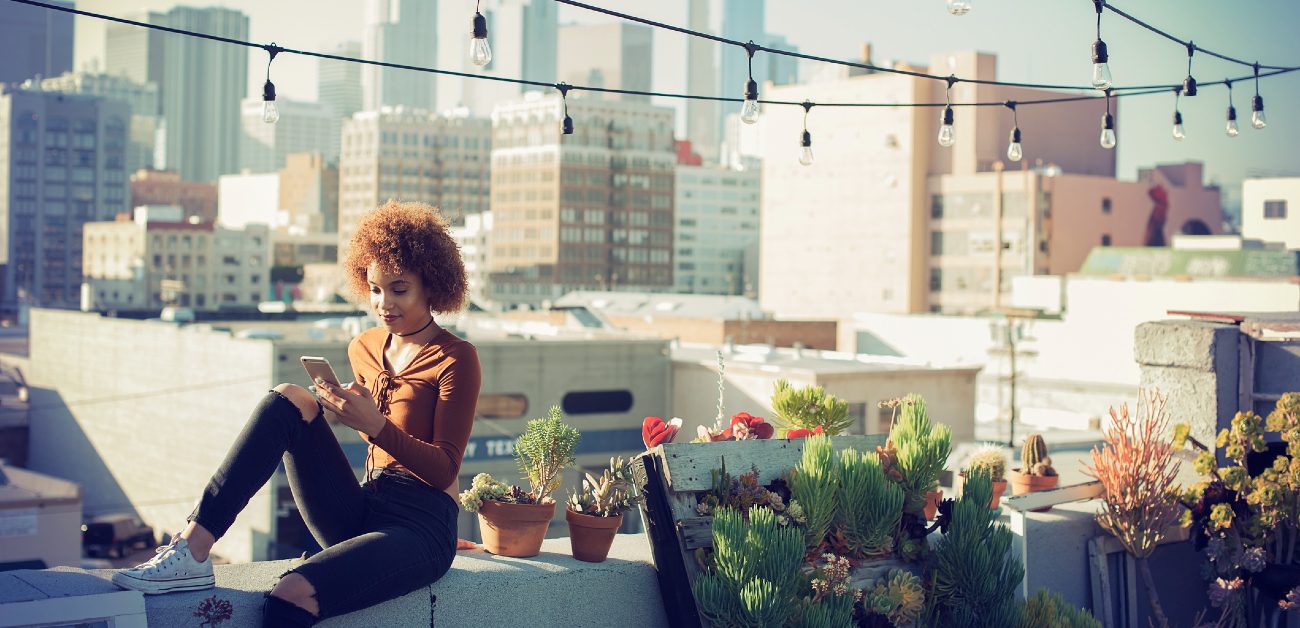 A woman sits on a city rooftop patio while using her phone, with buildings and skyline in the background.