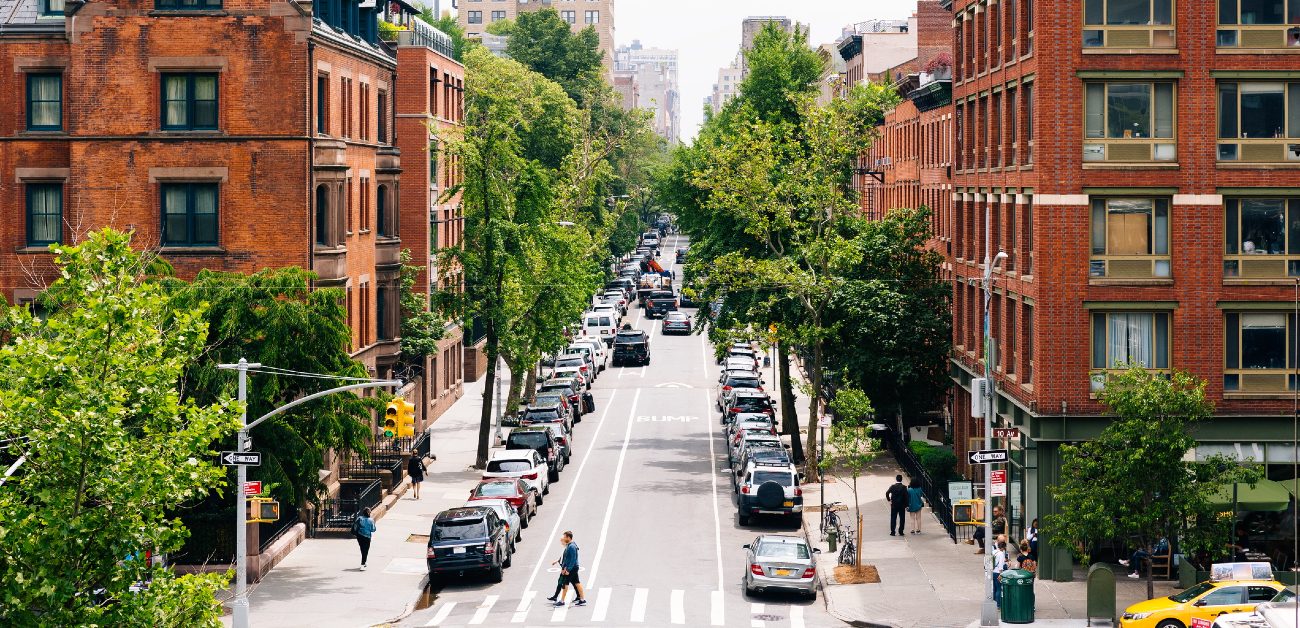 A picture looking down a city street filled with parked cars with trees and apartment buildings on each inside.