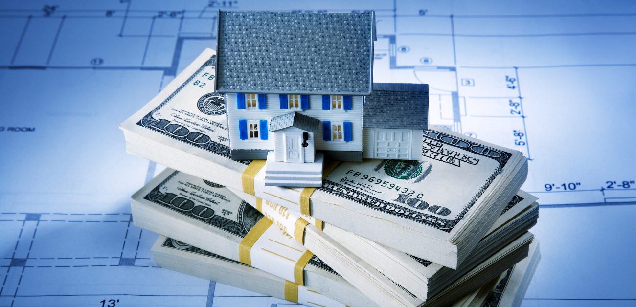 An image of a house miniature sitting upon three thick bound stacks of money sitting atop a blueprint.