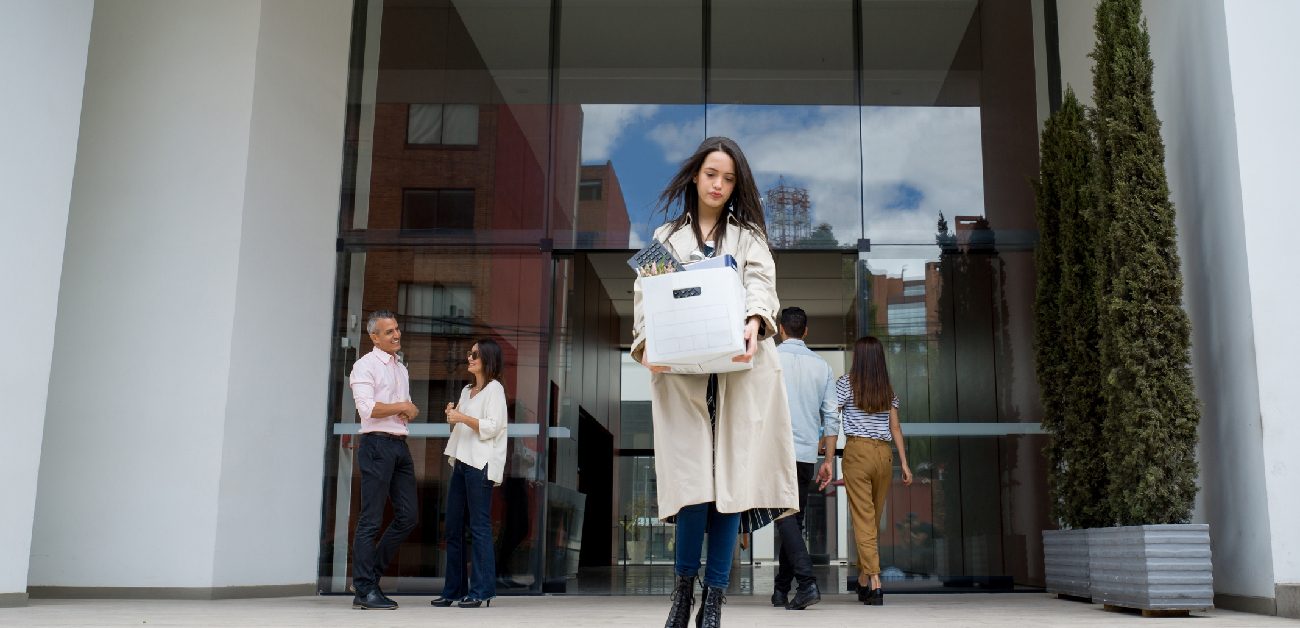 A woman in business attire leaving the front of an office building carrying personal items in a box.