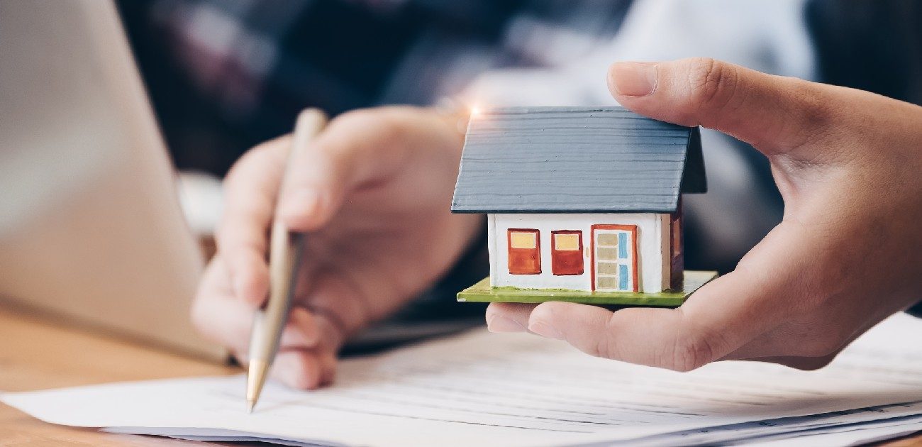 A picture of a person holding a house miniature and another person writing on a document with a pencil.