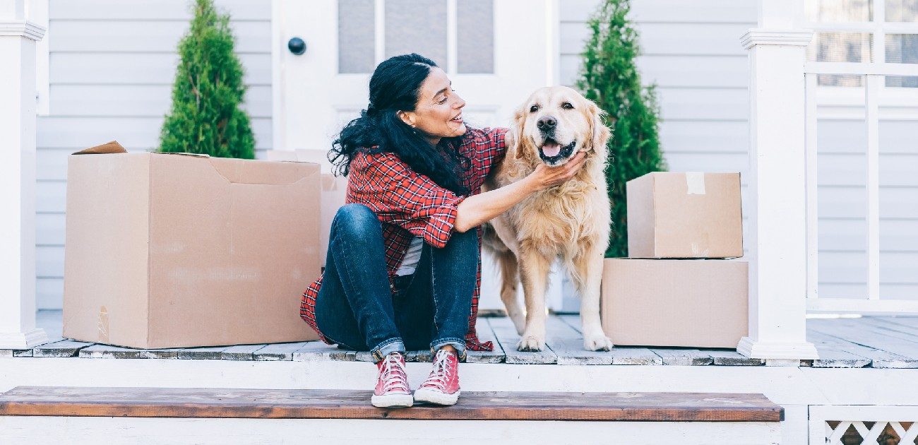 A woman sits with her dog on the front porch of a new home, with packing boxes around them.