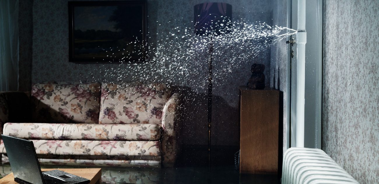 A photo of a living room with a burst pipe behind the wall spraying water from the right side across the room