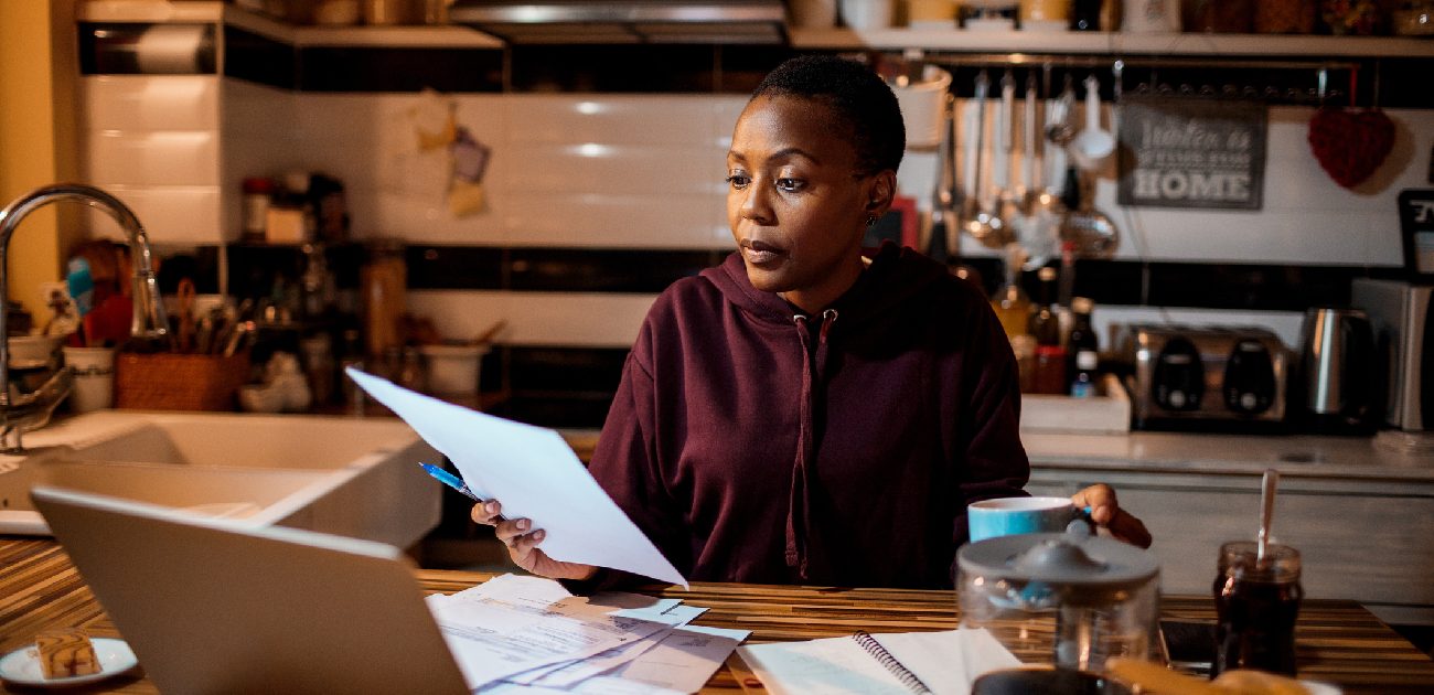 A woman sits at a kitchen table with a laptop while reviewing scattered bills and documents.