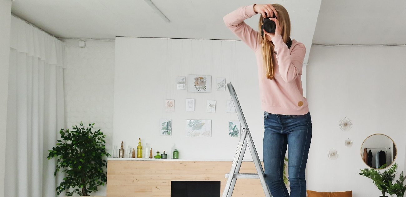 A woman photographer aims her camera toward the viewer while standing on a step stool.