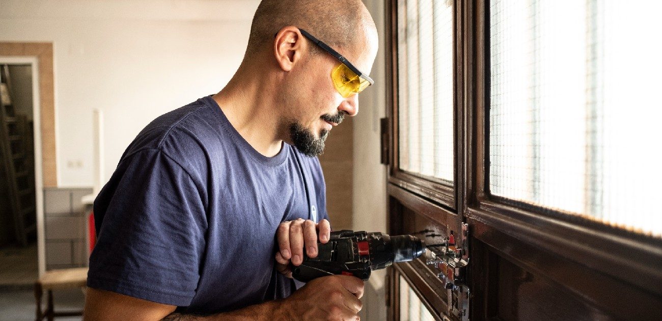 A man wearing safety glasses uses a power drill on a wall in his home.