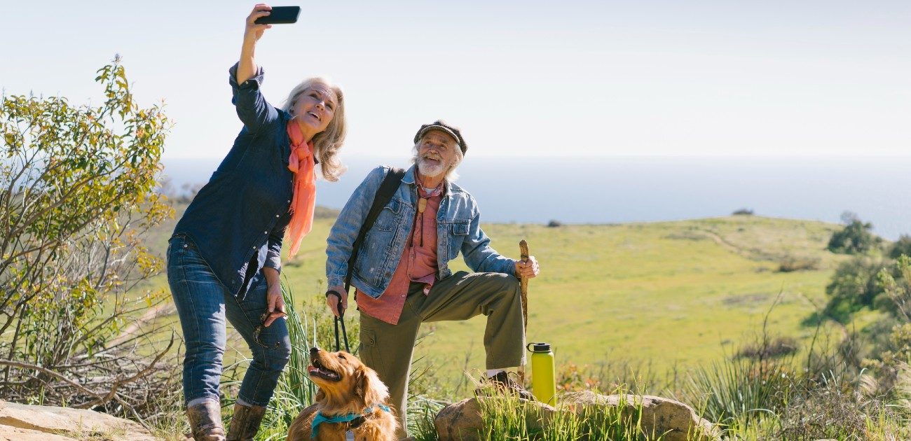 An elderly man and woman hiking take a selfie with their dog.