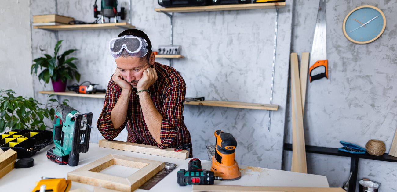 A man with his face resting on his hands standing at his work bench and looking stressed.