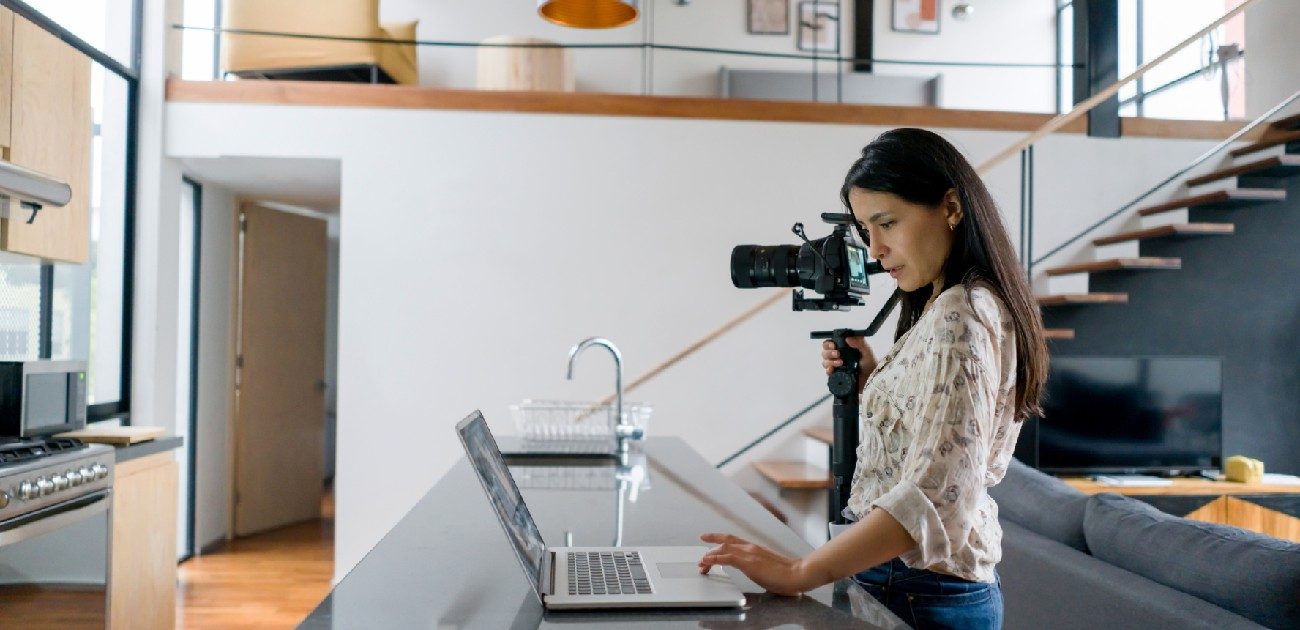 A woman working on a laptop at a tall table in a home with a video camera on a tripod next to her.