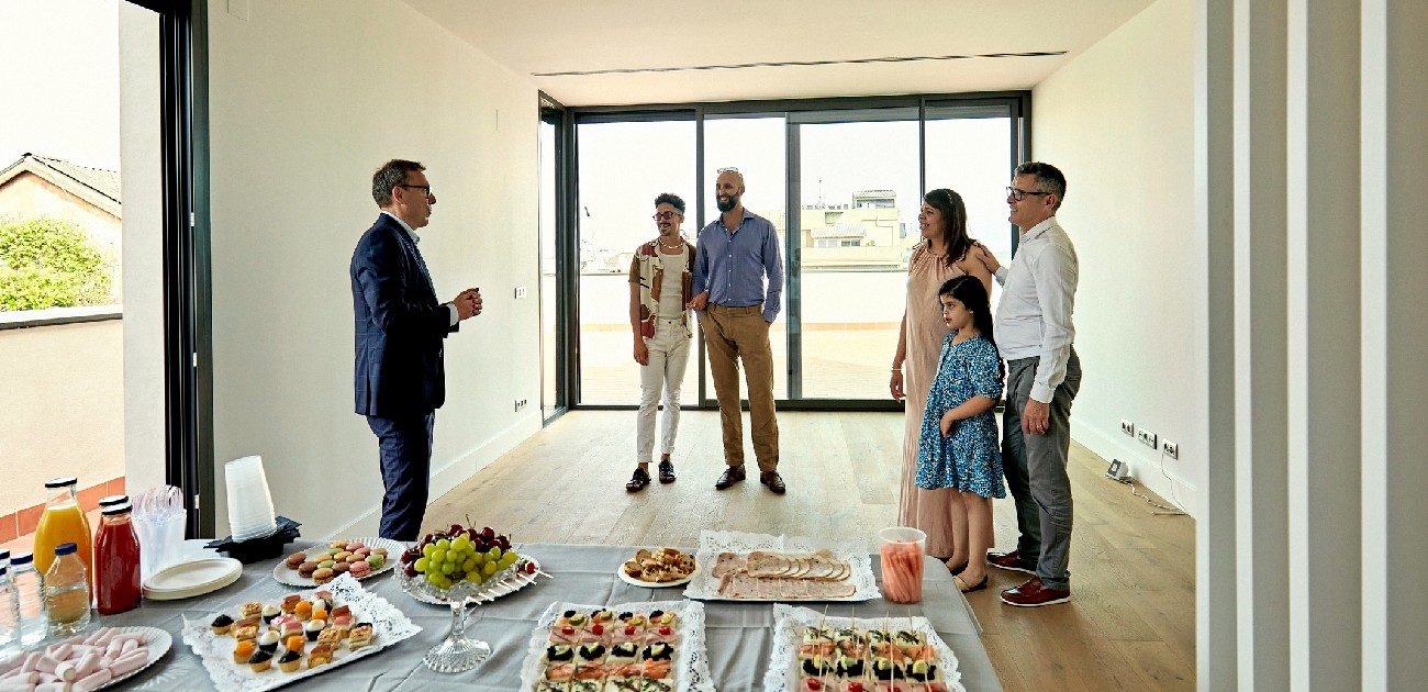 A family and a couple both tour an open house with the real estate agent present, and a table of assorted food and drink.