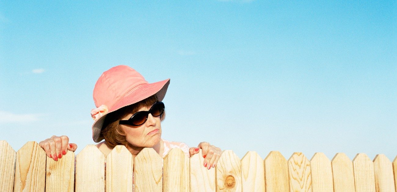 A picture of a woman in sunglasses and a hat peering over a fence with a disgruntled look on her face.