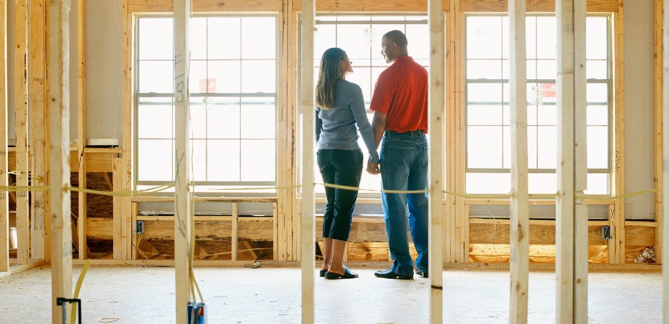 A man and a woman couple stand inside a home under construction, with only the wooden frame of the interior complete.