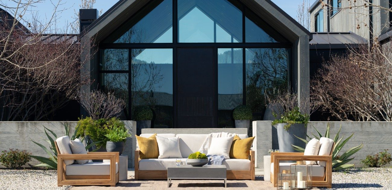 A picture of a newly built house with glass walls and patio furniture out in front of it.