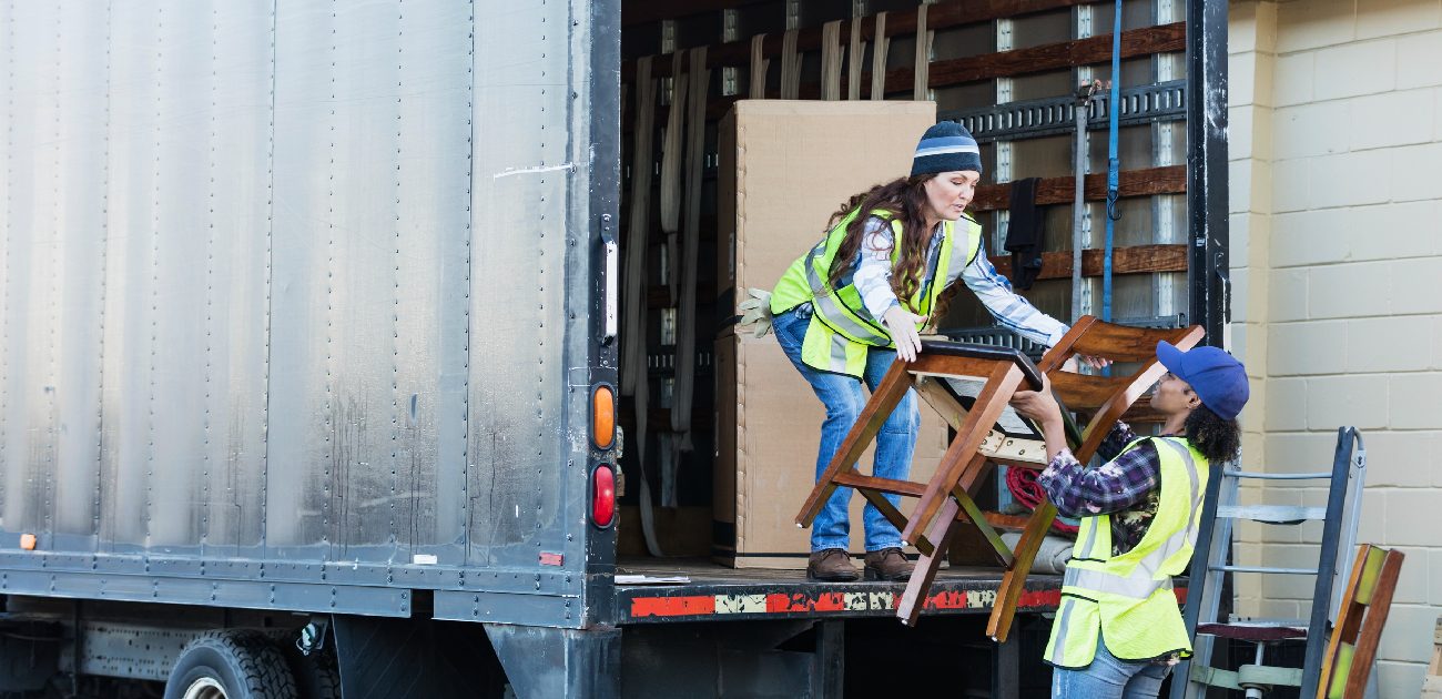 Two movers in safety vests lift a chair into the back of a loading truck.