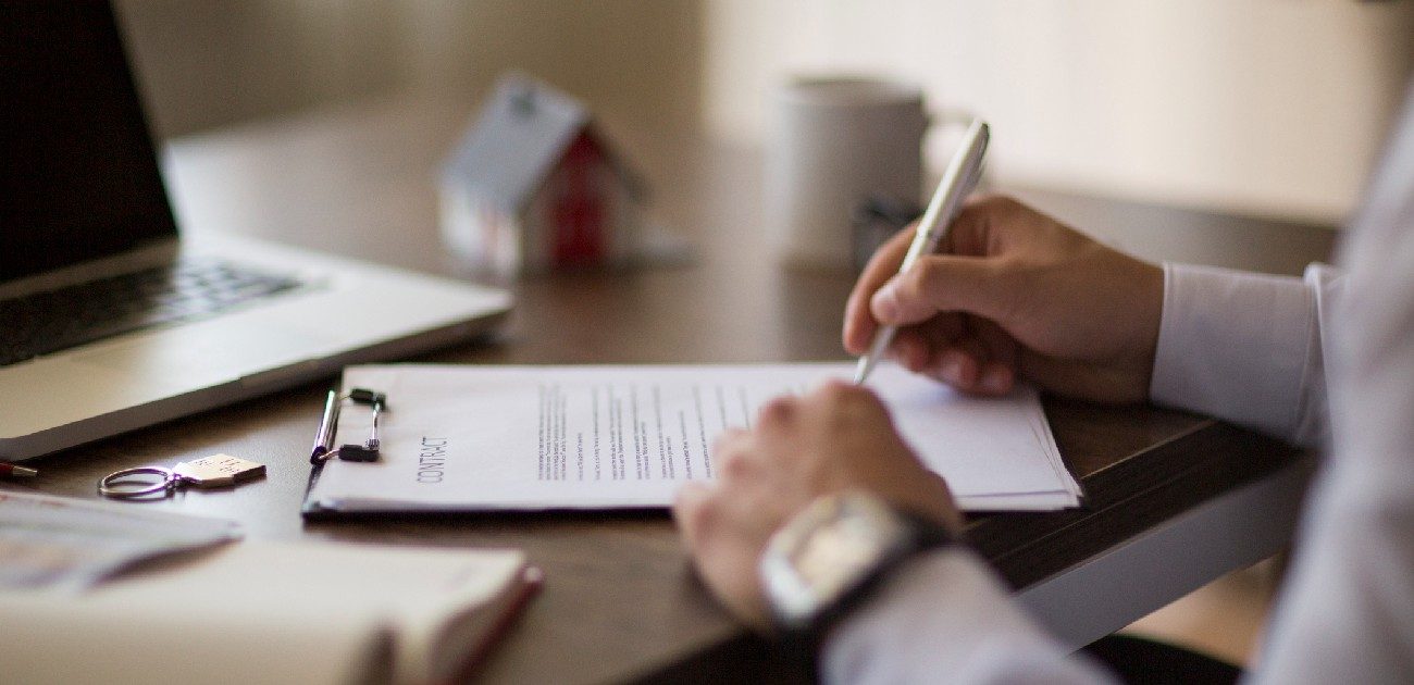 A picture of a person's hands signing a document on a clipboard at a desk.