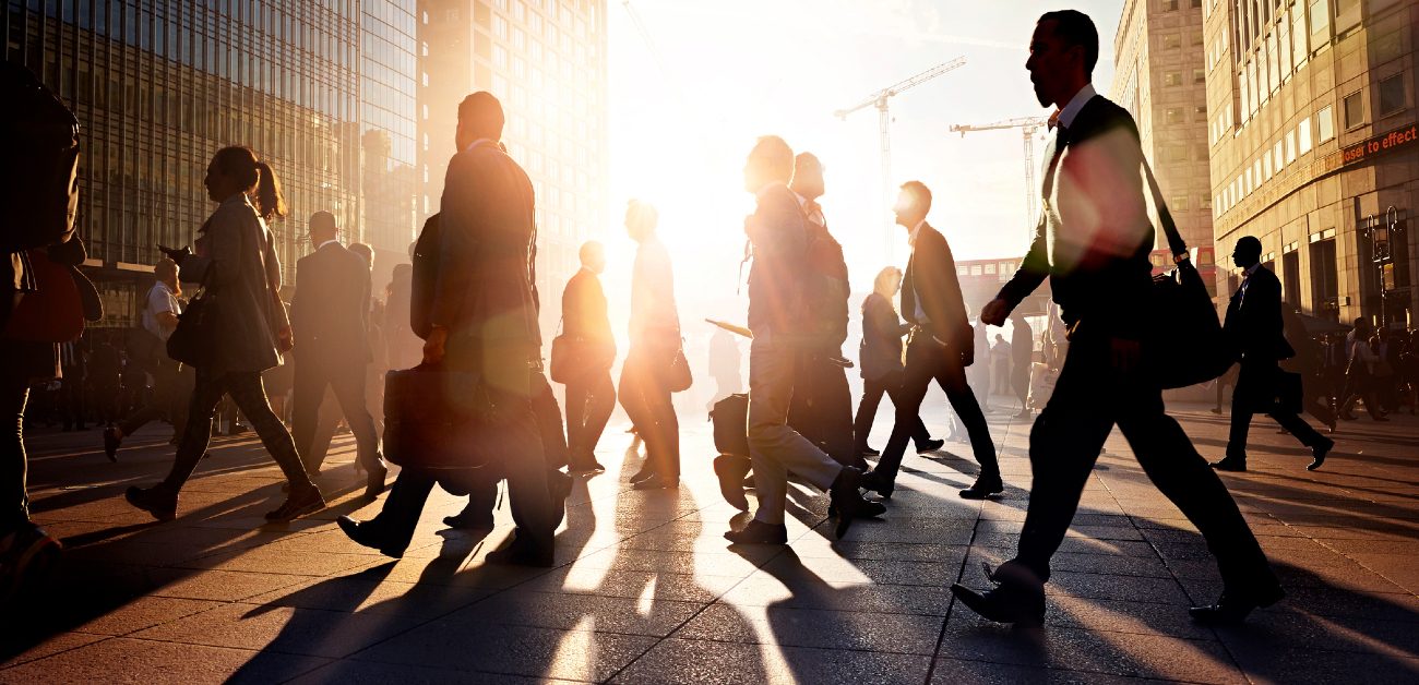A picture of morning commuters walking through a city in the morning, silhouetted by the early sunrise flare.