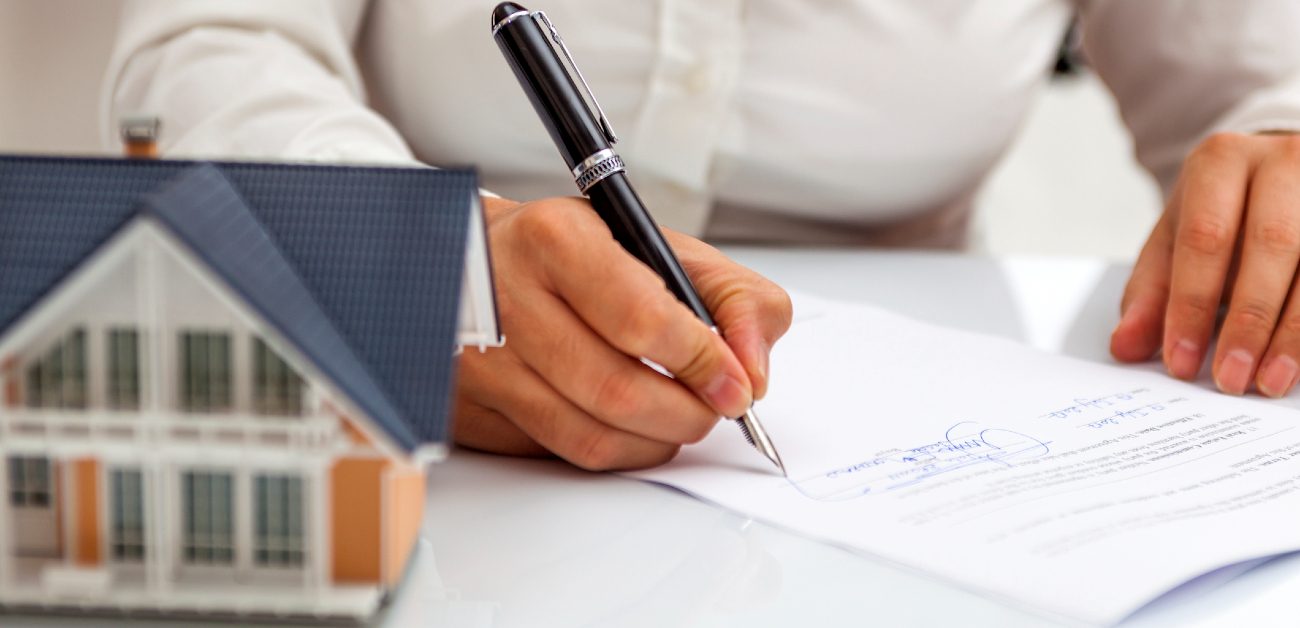 A picture of a person signing a document at a desk and a house miniature out of focus to the right of their signing hand..