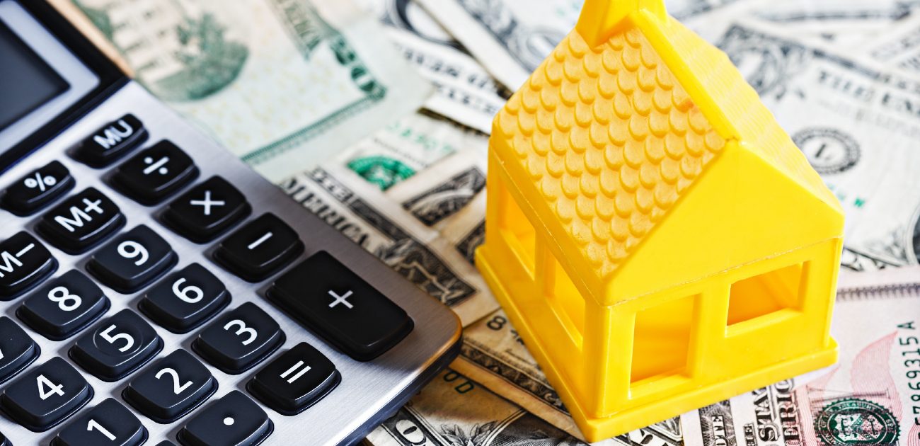 A yellow house miniature sits next to a calculator with scattered dollar bills underneath