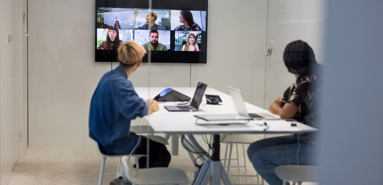 Two office workers in a conference room meet with the rest of their staff over a video call via the wall-mounted TV.
