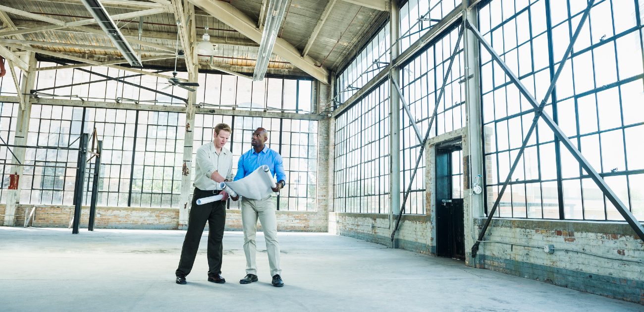 Two men in an empty commercial building look over blueprints and other construction plans.