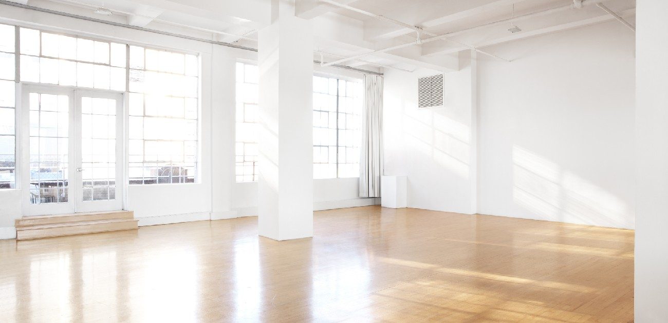 A picture of an empty room with hardwood floors and white walls, and bright daylight coming in from the windows.