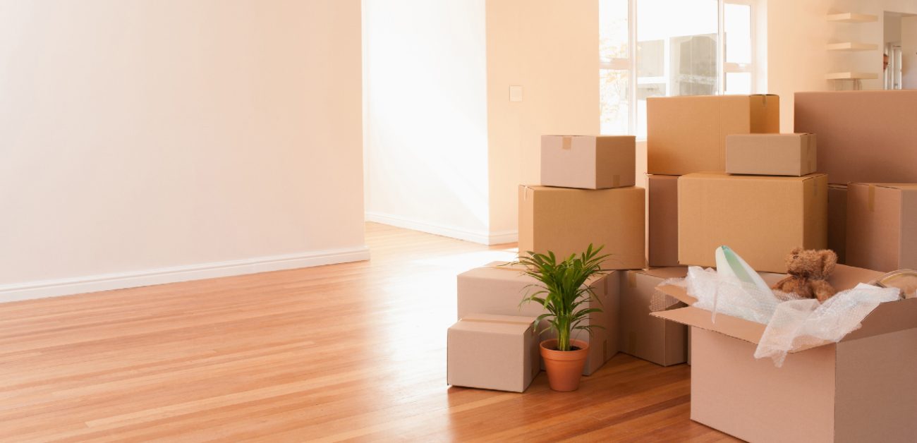 A picture of a bright, empty room with hardwood floors during the day, with stacks of boxes and belongings from a move.